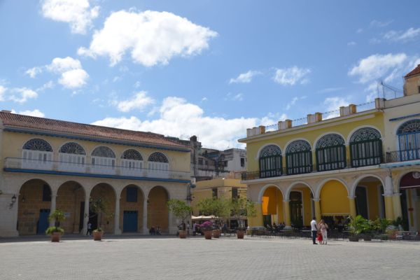 Auf dem Bild ist ein sonniger Platz in Havanna, Kuba zu sehen, umgeben von bunten Kolonialgebäuden mit Arkaden und Balkonen.