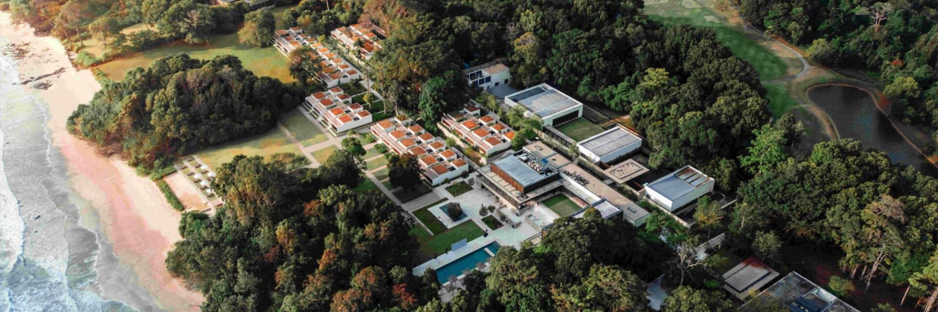 Luftaufnahme des Mandarin Oriental Desaru Coast Resorts in Malaysia, eingebettet zwischen tropischem Wald und Sandstrand am Meer. Die moderne Hotelanlage mit weißen Gebäuden und roten Dächern erstreckt sich entlang der Küste und wird von üppigem Grün umgeben.