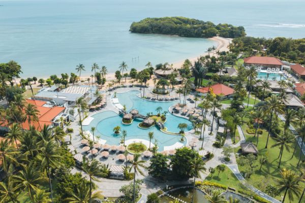 Das Foto zeigt eine weitläufige Hotelanlage auf Bali mit einem tropischen Garten, großen Palmen, einem geschwungenen Pool und direktem Zugang zum Sandstrand am Meer. Im Vordergrund sind zahlreiche Sonnenschirme und Liegen rund um den Pool zu sehen, während im Hintergrund das blaue Meer und eine kleine grüne Halbinsel ins Auge fallen.