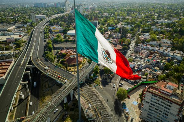 Luftaufnahme einer riesigen mexikanischen Flagge auf einem hohen Mast über einem Autobahnkreuz in Mexiko-Stadt, umgeben von dicht bebauten Wohnvierteln, Hochhäusern und grünen Hügeln im Hintergrund.