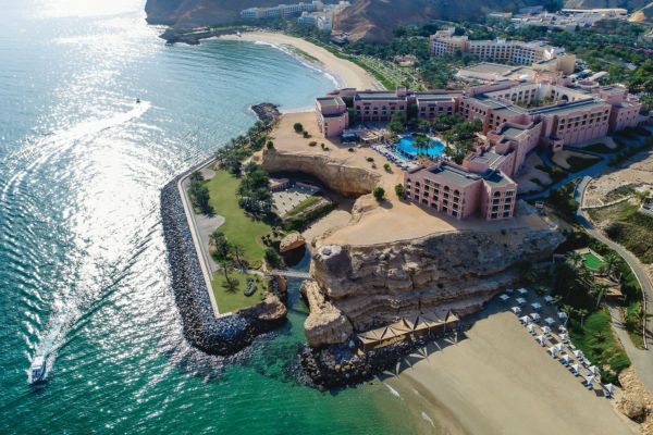 Luftaufnahme eines luxuriösen Hotels in Oman, das eindrucksvoll auf einer Klippe direkt am Meer liegt und von feinem Sandstrand sowie türkisblauem Wasser umgeben ist. Im Vordergrund fährt ein Boot über das sonnige Wasser, während die Hotelanlage mit Pools und Palmen einen exklusiven Urlaubsort präsentiert.