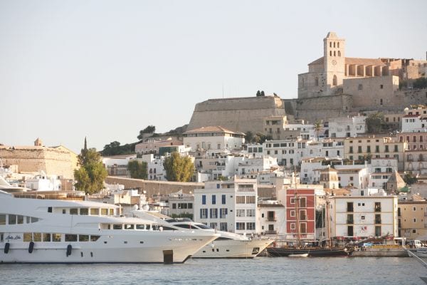 Im Vordergrund liegt eine luxuriöse weiße Yacht im Hafen von Ibiza, während im Hintergrund die Altstadt Dalt Vila mit historischen Gebäuden auf einem Hügel thront.
