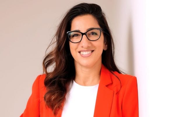 Auf diesem professionellen Business-Foto ist eine Frau mit langen braunen Haaren in einem leuchtend roten Blazer und weißem Oberteil vor einem neutralen Hintergrund zu sehen.
