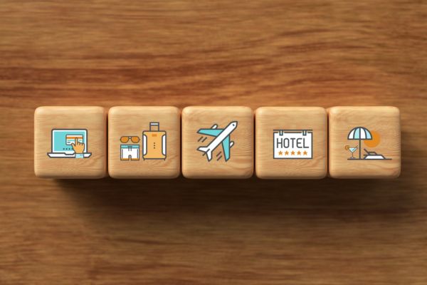 Fünf Holzklötze liegen nebeneinander auf einer Holzoberfläche, jeweils mit Reise-Icons bedruckt: Onlinebuchung, Reisebüro, Flugzeug, Hotel und Strandurlaub.