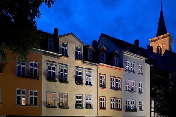 Mehrere pastellfarbene historische Häuser stehen dicht nebeneinander auf steinernen Bögen, die über einem beleuchteten Durchgang verlaufen. Die Fassaden sind gelb, orange und hellgrau, mit weißen Fenstern und Blumenkästen. Es ist Abend, der Himmel leuchtet tiefblau. Im Hintergrund ragt ein Kirchturm mit spitzem Dach empor. Die Szene wirkt ruhig und stimmungsvoll beleuchtet.