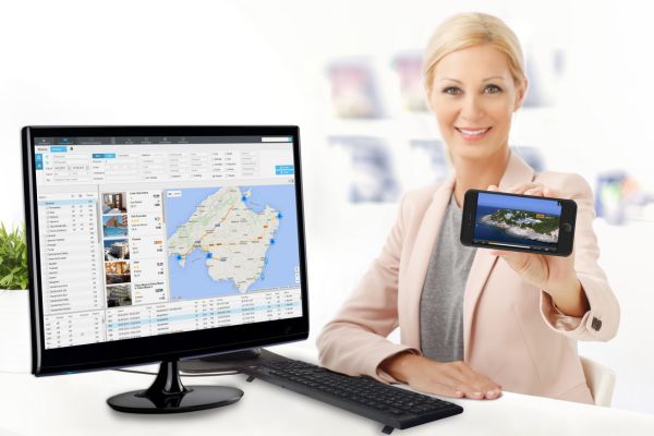 Business-Frau im hellen Büro sitzt an einem Schreibtisch mit Computer und Tastatur und hält ein Smartphone mit Urlaubsbild vor sich, auf dem Monitor ist eine Reisebuchungssoftware mit Karte und Hotelliste zu sehen.