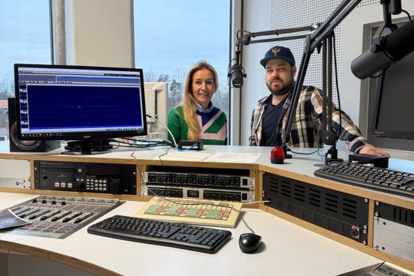 Zwei Personen sitzen in einem Radiostudio vor Mischpult, Monitoren und Mikrofonen und blicken in die Kamera.