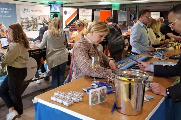 Auf dem Foto ist eine gut besuchte Informations‑ und Messestandsituation in einem Innenraum zu sehen, bei der mehrere Personen an verschiedenen Countern von Reise‑ und Touristikunternehmen miteinander sprechen, Prospekte betrachten und Werbegeschenke auf den Tischen liegen.