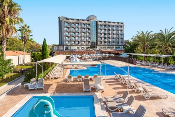 Großes Strandhotel mit mehreren Balkonen hinter einer weitläufigen Poollandschaft mit Liegestühlen, Sonnendächern und Kinderbecken mit blauem Elefantenrutschenmotiv, umgeben von Palmen und Gartenanlagen an einem sonnigen Tag.