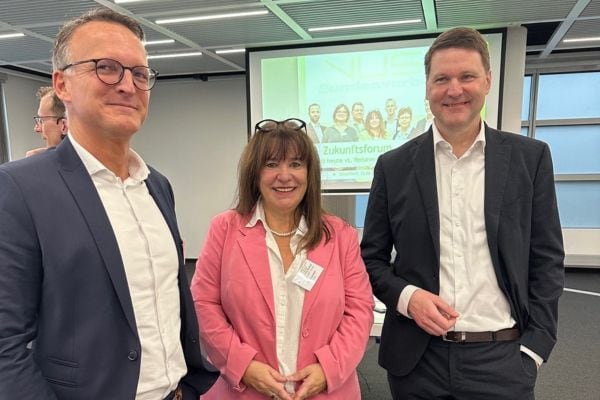 Drei Personen in Businesskleidung stehen in einem Konferenzraum nebeneinander und blicken in Richtung Kamera; im Hintergrund ist eine Präsentationsfolie mit dem Titel „Zukunftsforum“ sowie Teilnehmende auf einer Leinwand zu sehen.