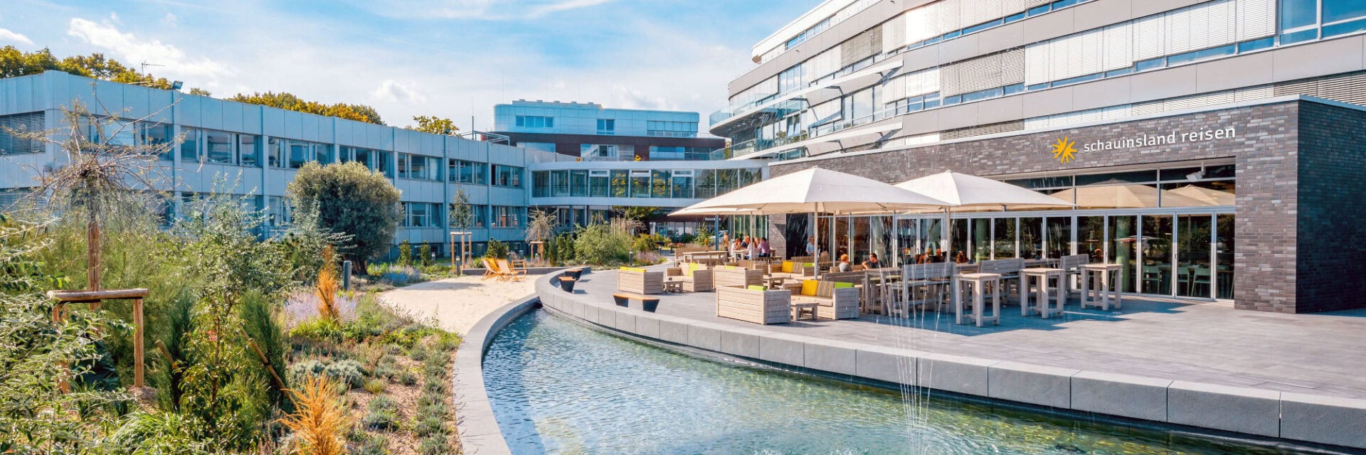 Das Bild zeigt das moderne Firmengebäude von Schauinsland-Reisen mit weitläufiger Terrasse, Sonnenschirmen und Sitzgelegenheiten am Wasserbecken. Im Vordergrund ist eine grüne Gartenlandschaft mit Bäumen und Sträuchern zu sehen.