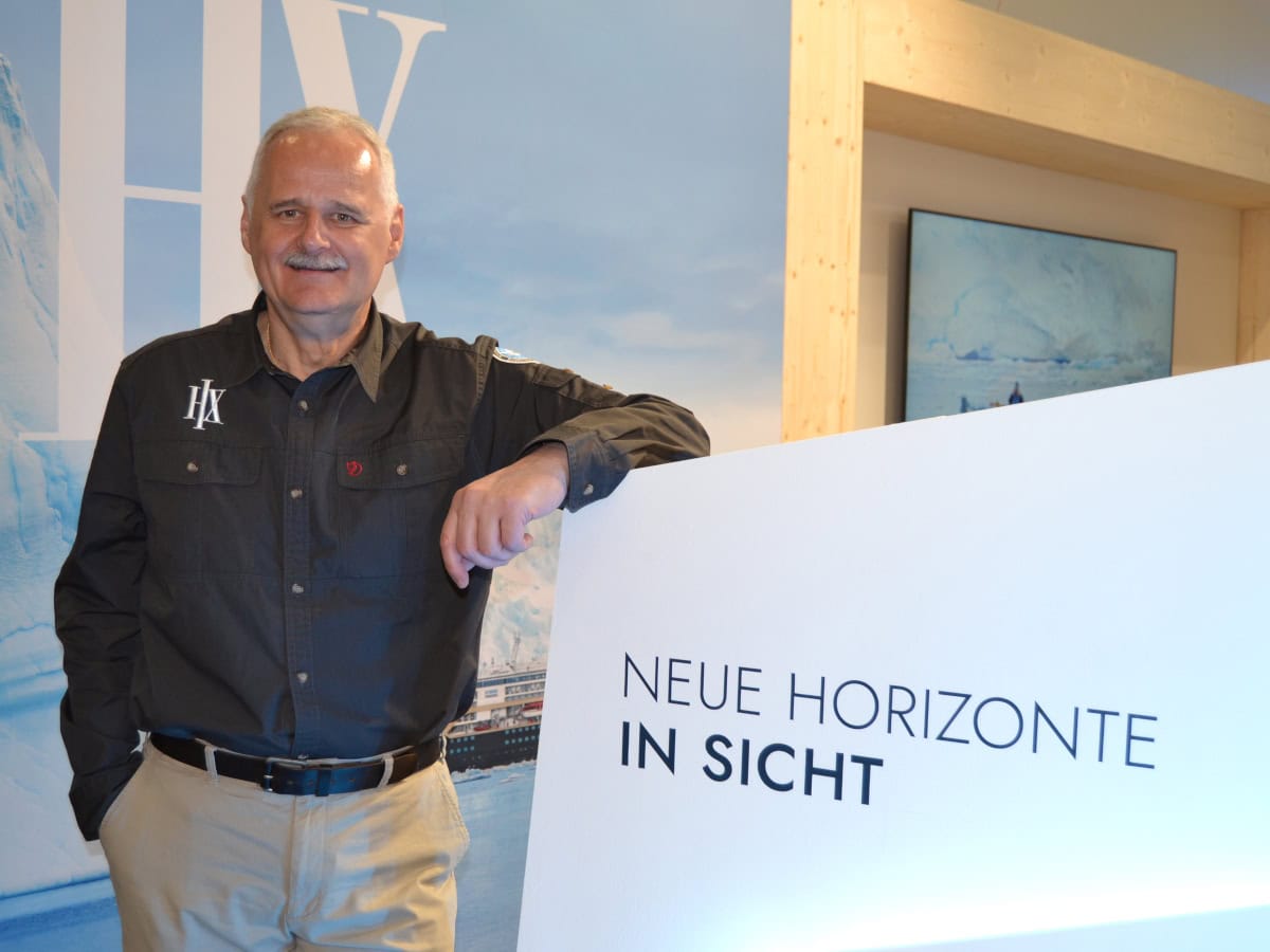 Ein Manager in schwarzem Hemd mit HX-Logo steht vor einem Messestand mit dem Slogan „Neue Horizonte in Sicht“, das Motiv zeigt einen modernen Messestand mit Arktis-Thema und einem Bildschirm im Hintergrund.