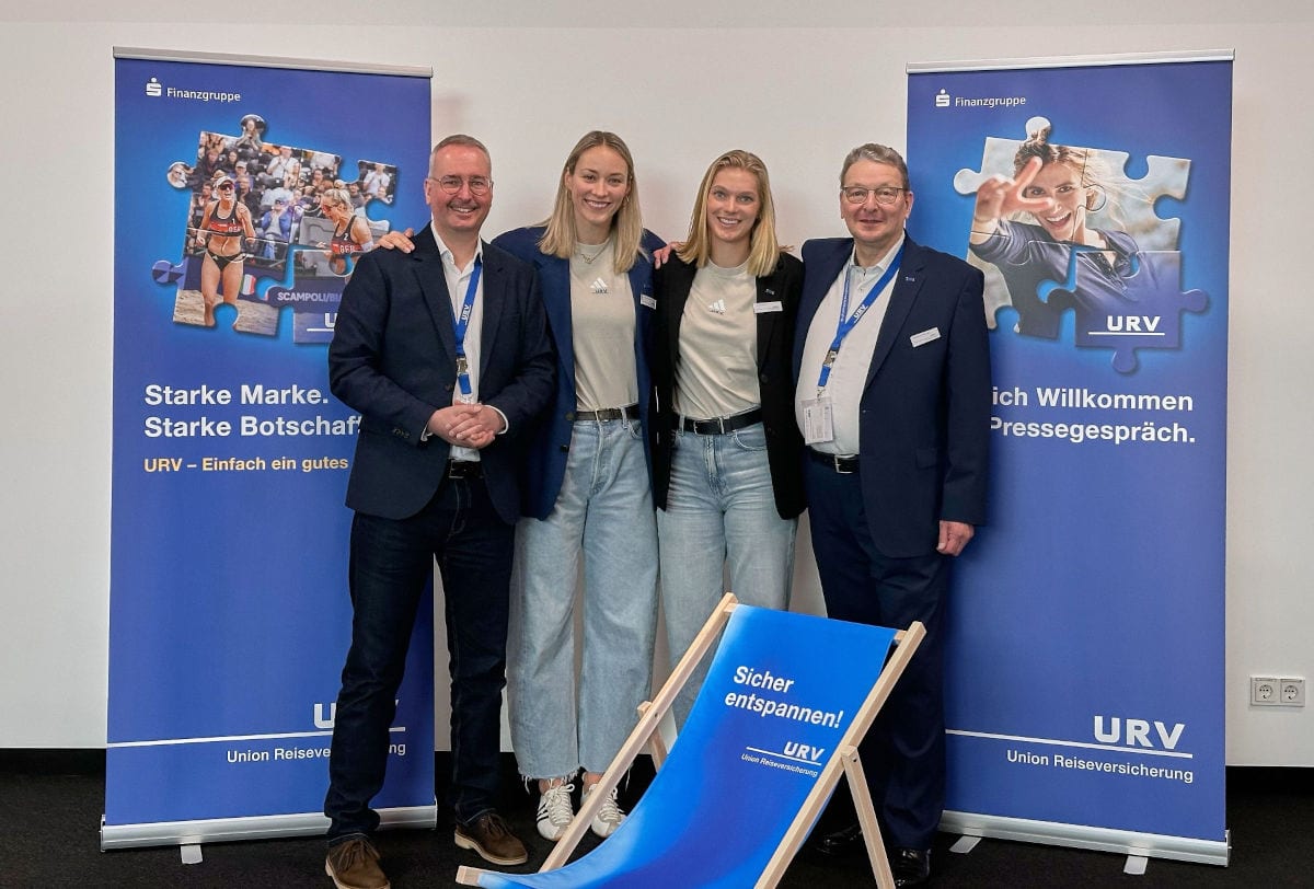 Vier Personen stehen nebeneinander vor zwei blauen Roll-up-Bannern der URV/Finanzgruppe. Zwei Männer im Anzug mit Namensschildern werden von zwei sportlich gekleideten Frauen flankiert. Davor steht ein blauer Liegestuhl mit der Aufschrift „Sicher entspannen! URV“.