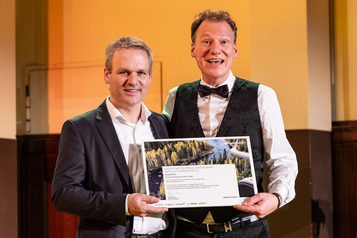 Zwei Männer stehen nebeneinander auf einer Bühne mit warm beleuchtetem Hintergrund und tragen Headset-Mikrofone. Der Mann links trägt einen dunklen Anzug mit weißem Hemd, der Mann rechts ein weißes Hemd mit schwarzer Weste und Fliege. Gemeinsam halten sie eine gerahmte Urkunde mit der Aufschrift „Certificate of Green Partnership with Lufthansa Group“, versehen mit einem Landschaftsbild und mehreren Airline-Logos. Beide lächeln in die Kamera.