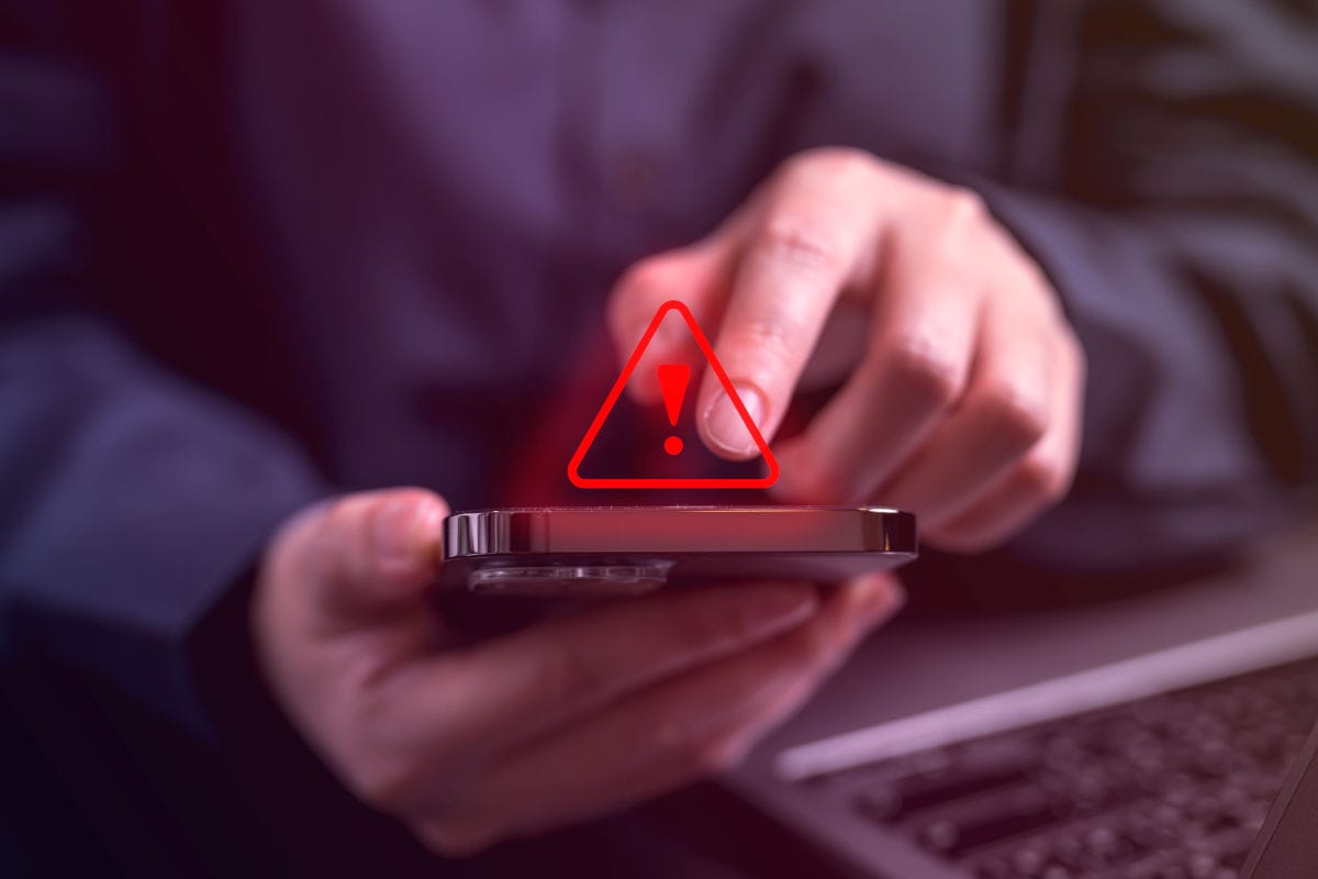Eine Person hält ein Smartphone in der Hand und tippt darauf, während ein rotes Warnsymbol mit Ausrufezeichen auf dem Display erscheint – das Bild thematisiert Online-Betrug, Handy-Sicherheit und Gefahren bei Flugbuchungen. Im Hintergrund ist unscharf ein Laptop zu erkennen, was auf digitale Risiken im Internet hinweist.