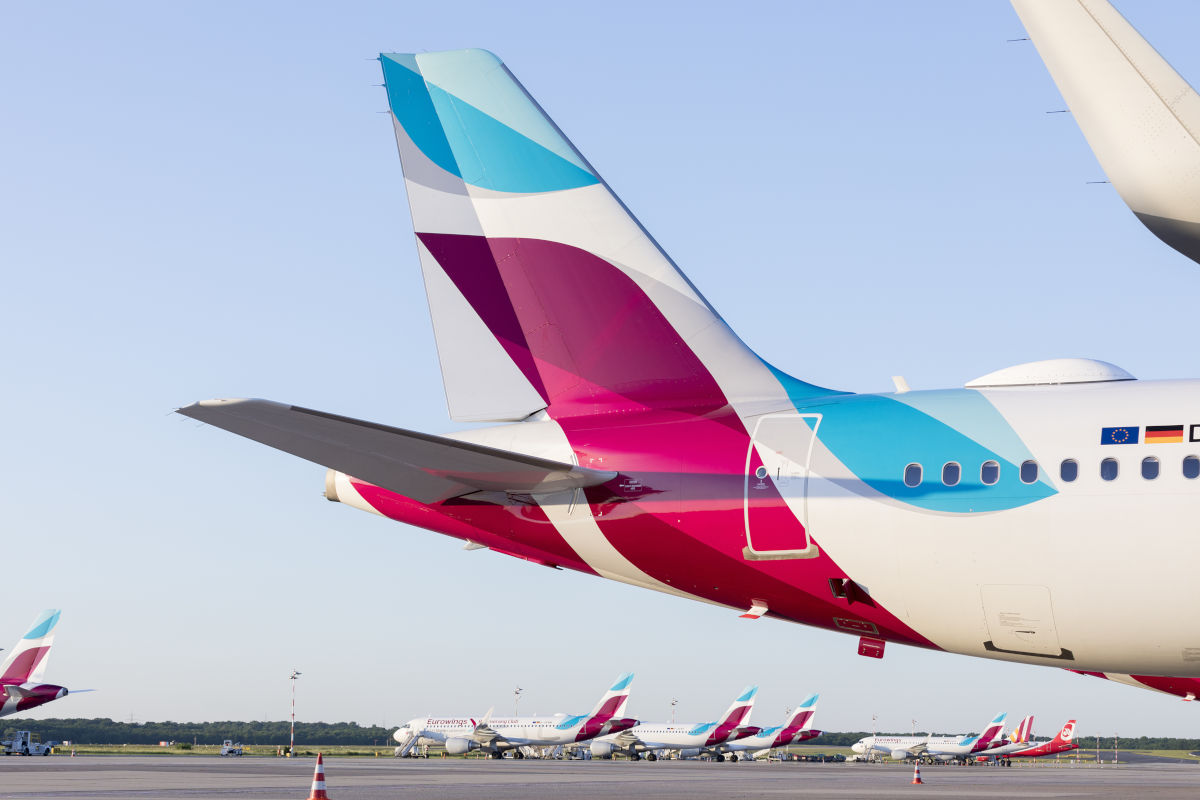 Auf diesem Foto ist das Leitwerk eines Eurowings-Flugzeugs auf dem Rollfeld eines Flughafens zu sehen, im Hintergrund stehen weitere Flugzeuge der Airline. Das Bild zeigt die markante blau-pinke Lackierung und die moderne Flugzeugflotte von Eurowings.