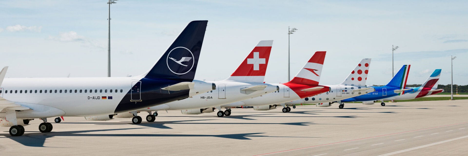 Auf dem Foto sind Flugzeuge mehrerer Airlines der Lufthansa Group wie Lufthansa, Swiss und Austrian Airlines auf einem Flughafen zu sehen, die deutlich ihre jeweiligen Logos zeigen. Die Maschinen stehen nebeneinander auf dem Vorfeld.