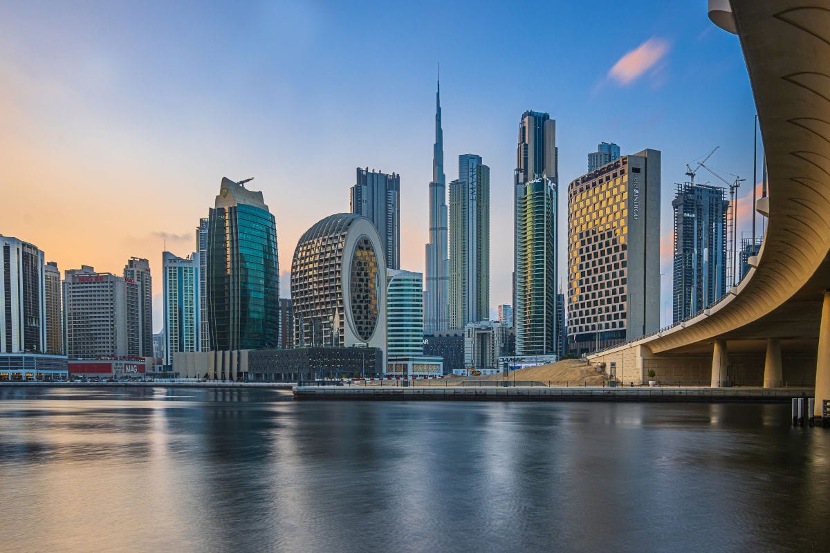 Skyline von Dubai am Wasser bei Sonnenuntergang mit dem Burj Khalifa im Zentrum, umgeben von modernen Hochhäusern und einer geschwungenen Brücke im Vordergrund.