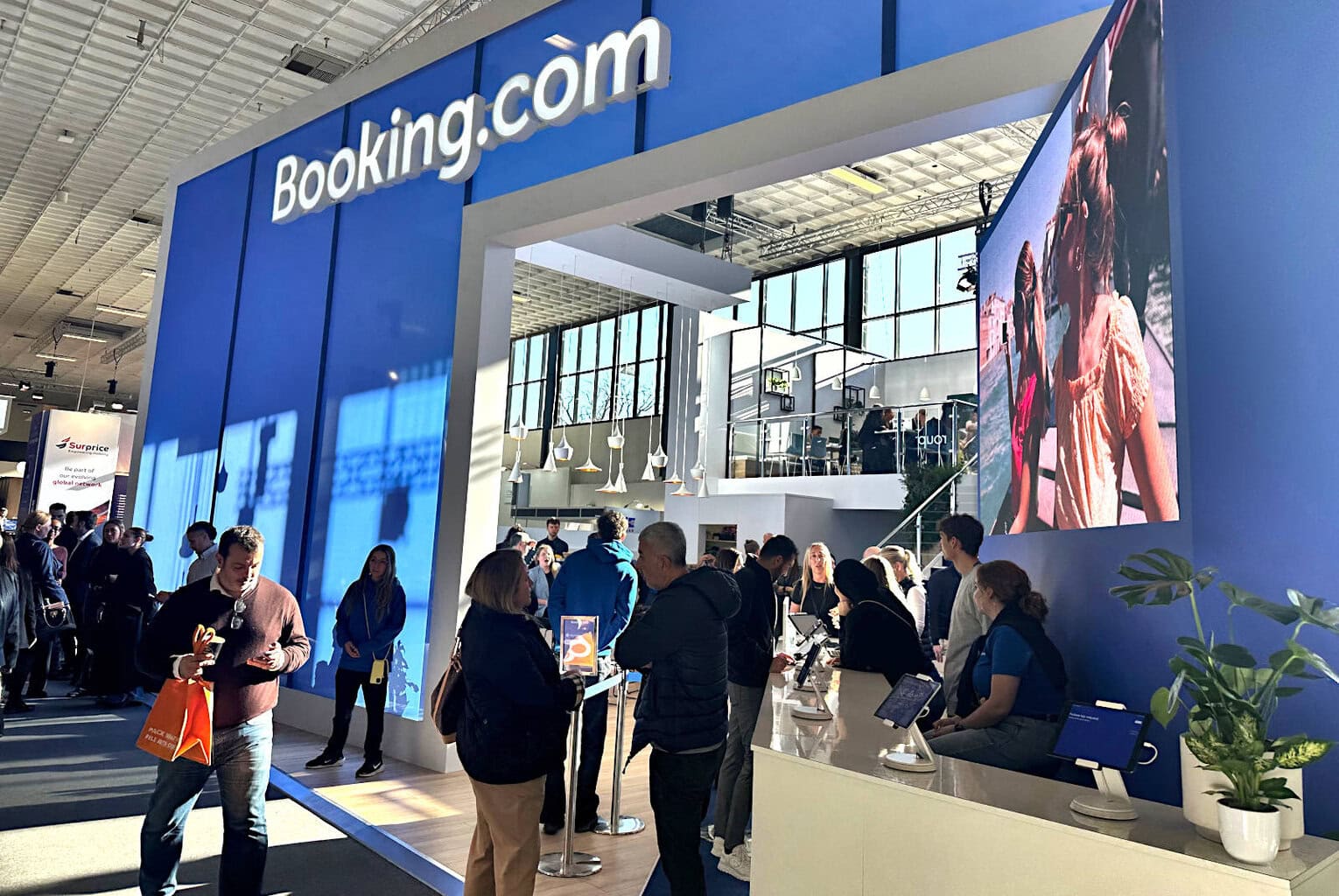 Großer Messestand von Booking.com auf der Reisemesse ITB in Berlin, mit blauem Markenauftritt, großer Videowand mit Urlaubsszene und vielen Besucherinnen und Besuchern, die sich an Countern informieren.