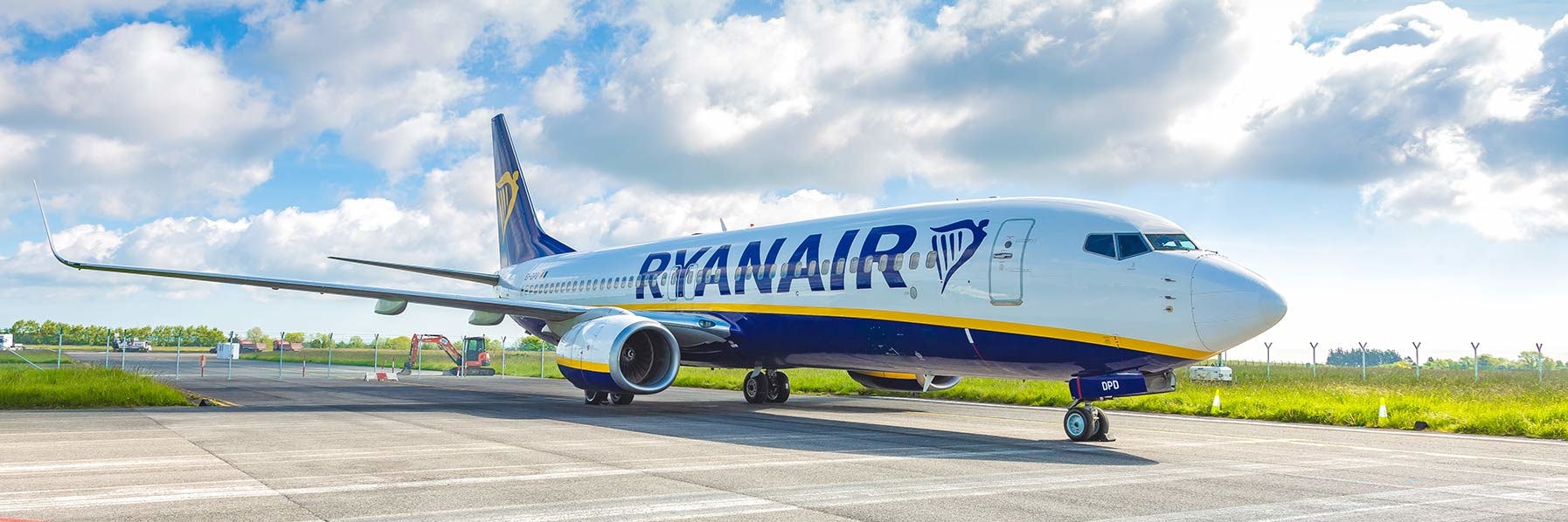 Auf dem Foto ist ein Ryanair-Flugzeug auf dem Vorfeld eines Flughafens bei Tageslicht zu sehen, die Maschine steht bereit zur Abfertigung. Im Hintergrund sind blauer Himmel mit einigen Wolken, grüne Wiese und Flughafenfahrzeuge sichtbar.