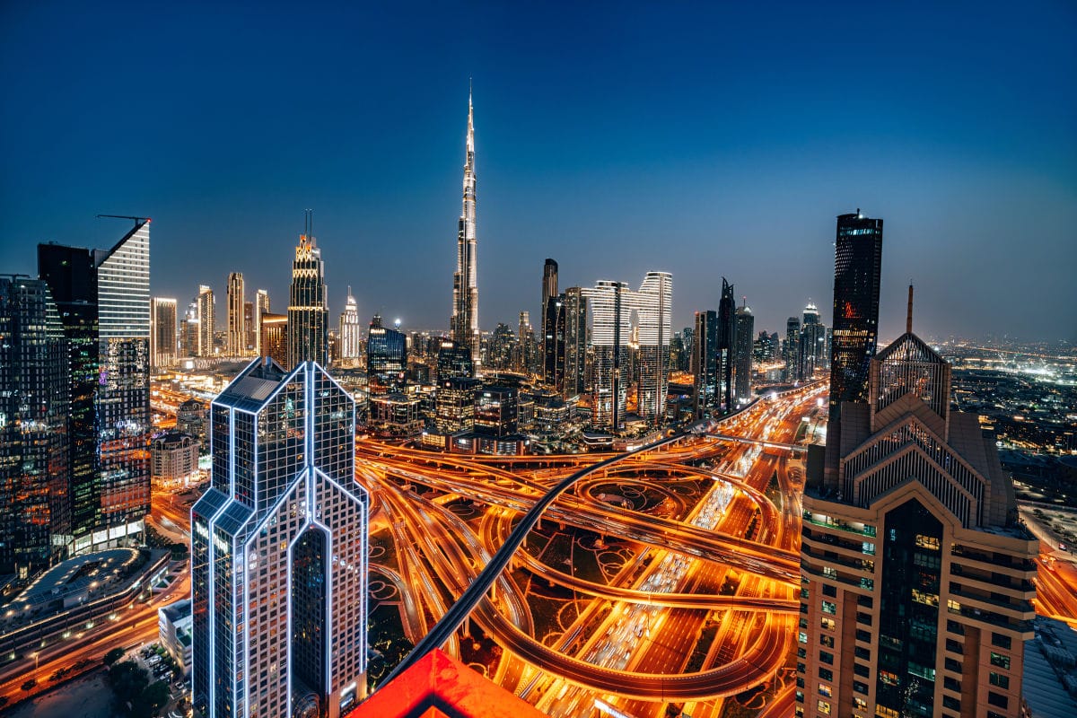 Nächtliche Skyline einer modernen Großstadt mit vielen beleuchteten Wolkenkratzern und einem großen Autobahnkreuz mit dichtem Verkehr; in der Mitte ragt ein sehr hoher Turm in den Himmel.