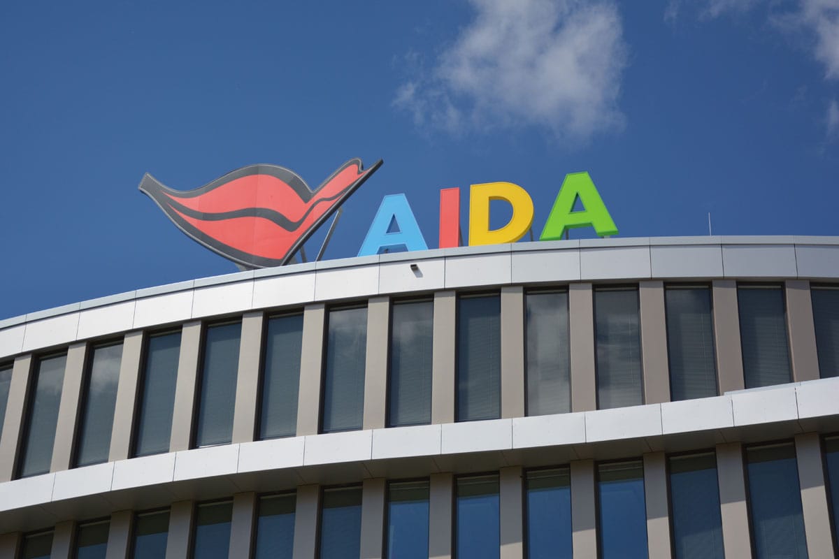 Die Aufnahme zeigt die moderne Fassade der AIDA Zentrale mit dem auffälligen, bunten AIDA-Logo und dem roten Kussmund auf dem Dach des Bürogebäudes vor strahlend blauem Himmel.