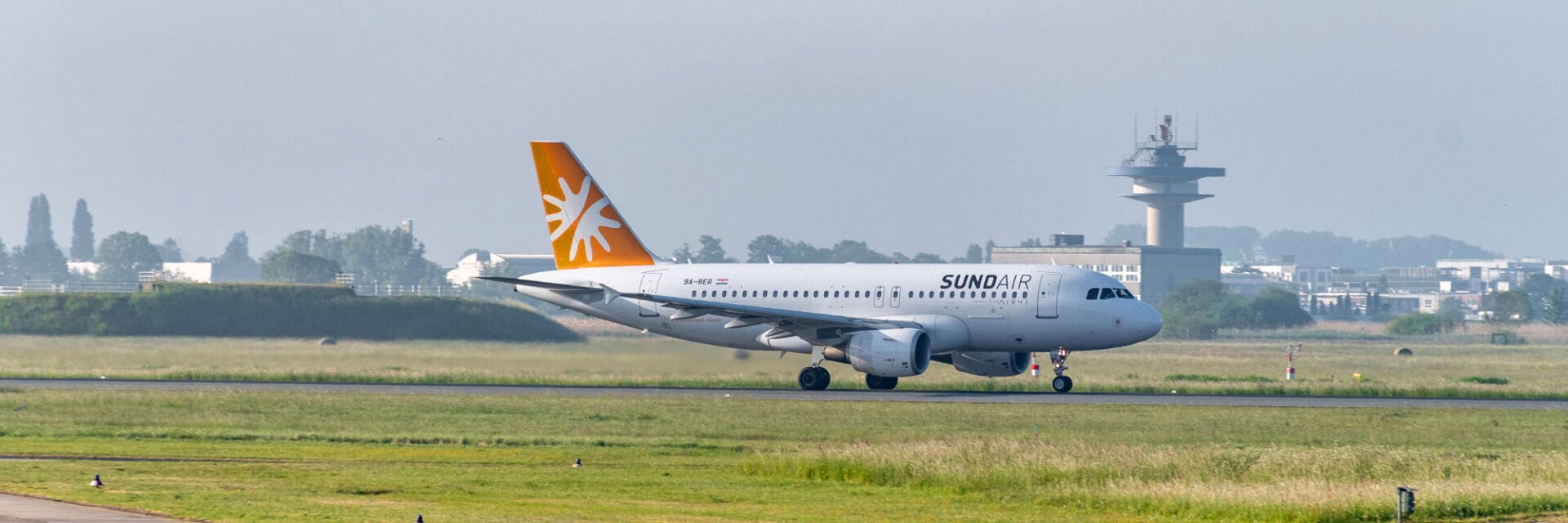 Auf dem Bild ist ein Sundair-Flugzeug auf der Startbahn des Flughafens Bremen zu sehen, im Hintergrund erkennt man den markanten Tower und einige Gebäude. Das weiße Flugzeug mit orangefarbenem Leitwerk steht bereit zum Start, umgeben von grüner Wiese und leichtem Morgennebel.