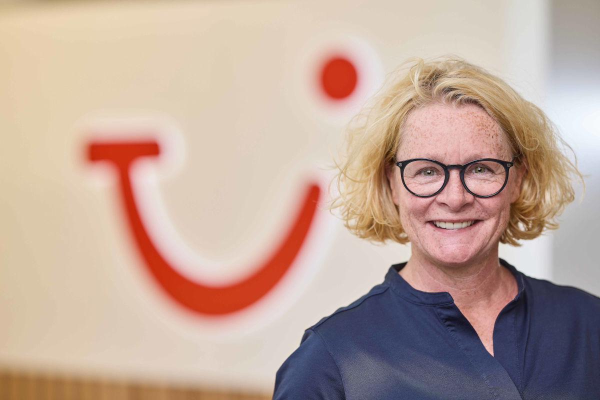 Lächelnde blonde Frau mit halblangen Haaren und dunkelgerahmter Brille vor einem Logo des Reiseveranstalters TUI
