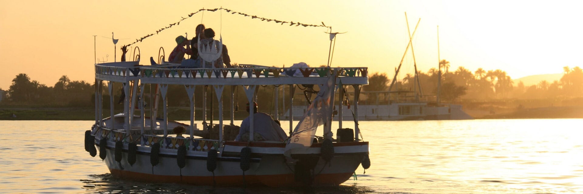 Boot mit mehreren Personen bei Sonnenuntergang auf dem Nil vor der Küste von Luxor, Ägypten, mit warmem goldenen Licht auf dem Wasser und Palmen sowie weiteren Booten im Hintergrund.