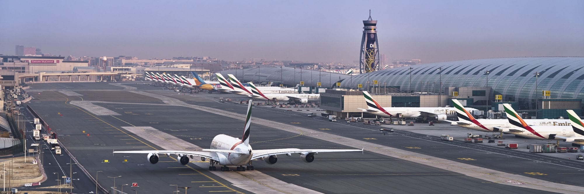 Das Bild zeigt das Vorfeld des Dubai International Airport mit mehreren Emirates-Flugzeugen, die nebeneinander vor dem modernen Terminalgebäude geparkt sind. Im Vordergrund rollt ein Airbus A380 der Fluggesellschaft Emirates über das Rollfeld.