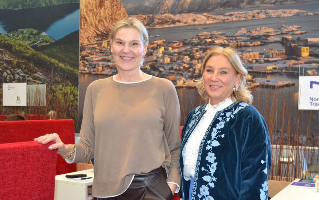 Zwei Reedereivertreterinnen stehen auf einem Reisemessestand, im Hintergrund ist ein großes Bild mit einer Norwegen-Aufnahme zu sehen.