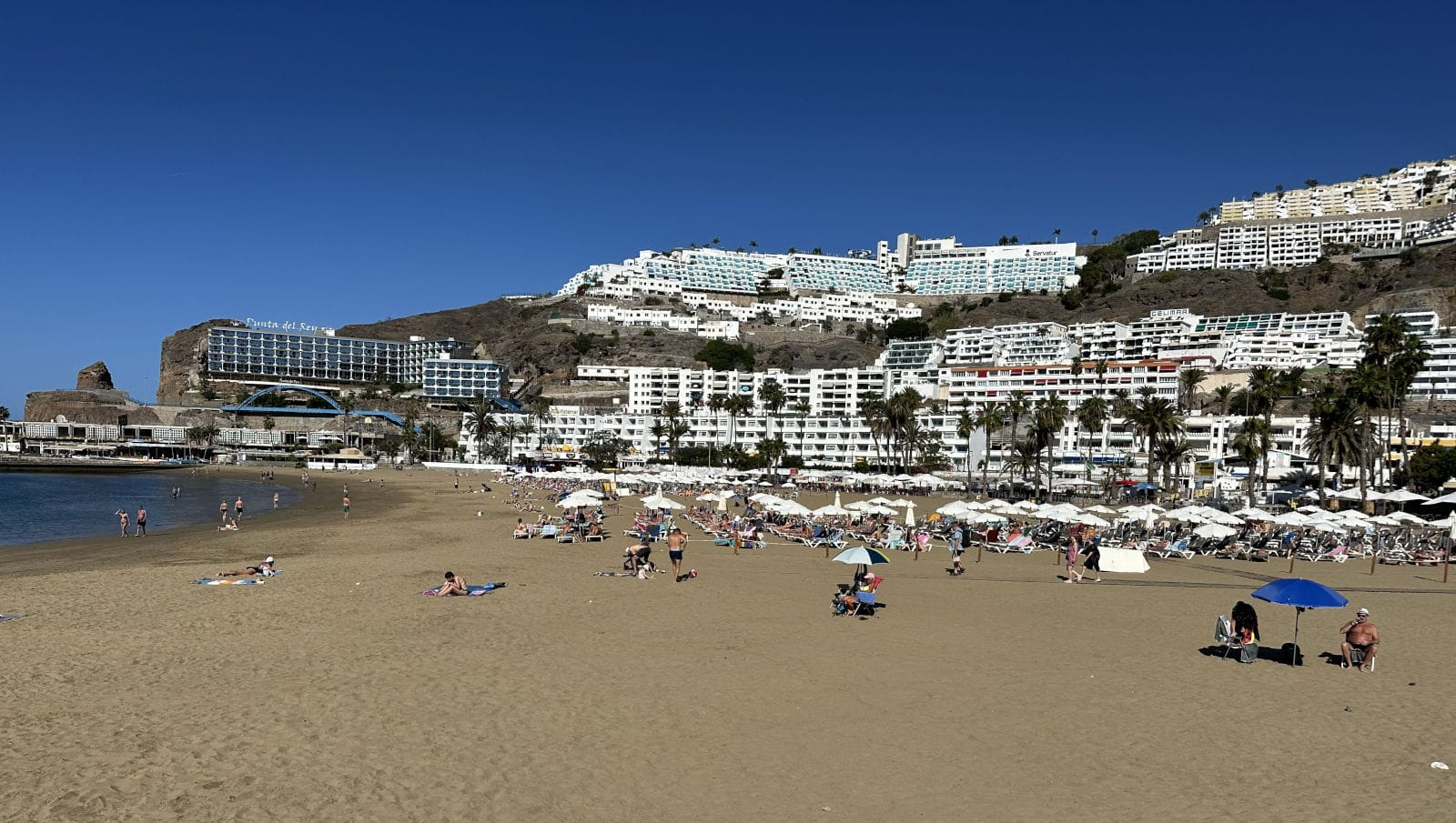 Strandurlaubsszene in Playa del Inglés, Maspalomas auf Gran Canaria, Spanien: Golden-gelber Sandstrand mit sonnenden Badegästen, bunten Sonnenschirmen und Liegen. Im Hintergrund erstreckt sich unter klarem blauem Himmel eine malerische Ansammlung weißer Hotels und Apartmenthäuser die steile Hügel hinauf bis zum Meer, mit Palmen und Felsenakzenten.