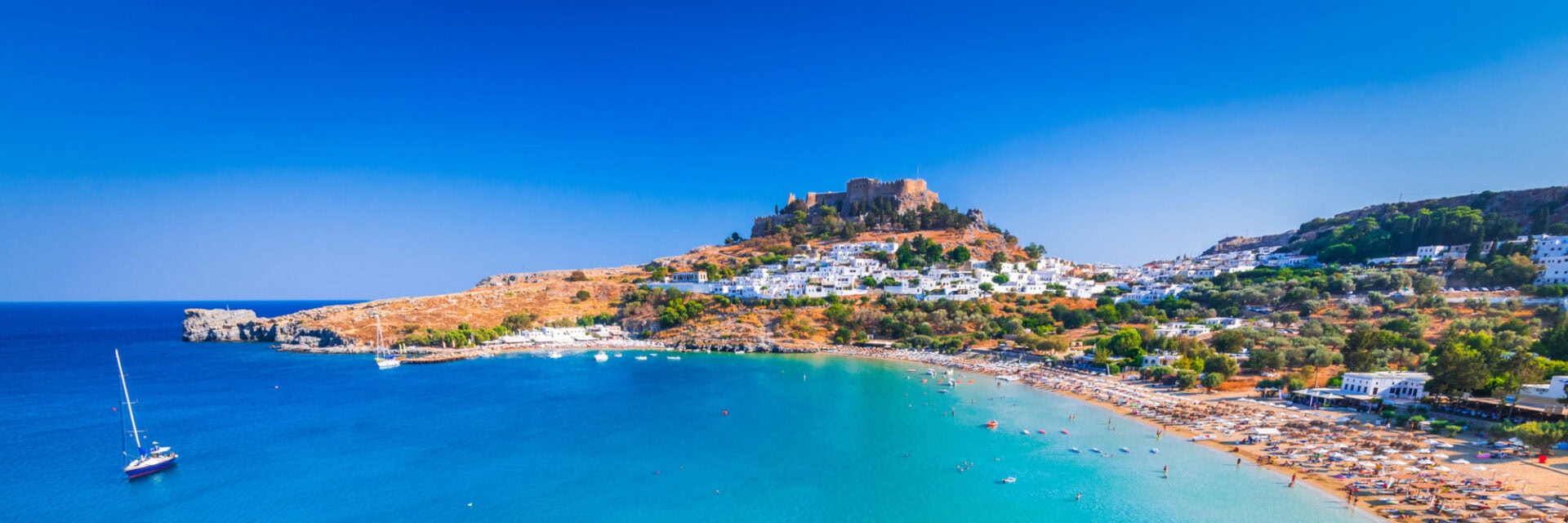 Weite Küstenbucht mit türkisblauem Wasser, Sandstrand und vielen Sonnenschirmen; im Hintergrund ein Ort mit weißen Häusern und einer Burg auf einem Hügel, im Vordergrund felsige Landschaft.