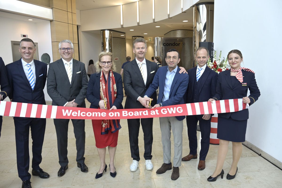 Bei der feierlichen Eröffnung der neuen Condor-Zentrale stehen mehrere Personen in Businesskleidung in einer modernen Empfangshalle und halten gemeinsam ein rot-weißes Band mit der Aufschrift „Welcome on Board at GWG“. Im Hintergrund ist das Condor-Logo sichtbar, während die Beteiligten zur symbolischen Banddurchtrennung goldene Scheren bereithalten. Das Bild illustriert einen wichtigen Moment für die Einweihung eines Firmengebäudes und den Unternehmensstart.