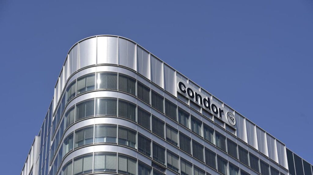 Das Foto zeigt die moderne Zentrale von Condor Airlines mit glänzender Glasfassade und großem Condor-Logo vor blauem Himmel. Das Bürogebäude hebt sich architektonisch durch abgerundete Ecken und viele Fenster hervor.