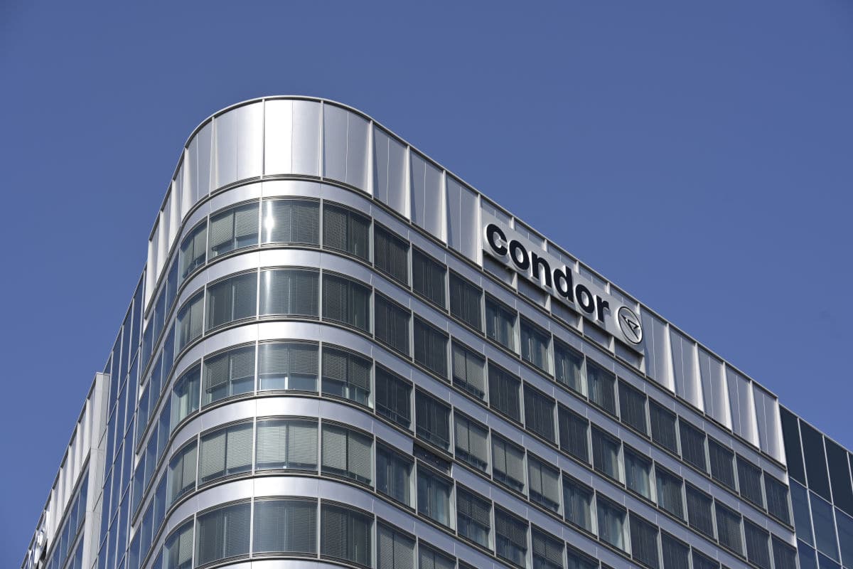 Das Foto zeigt die moderne Zentrale von Condor Airlines mit glänzender Glasfassade und großem Condor-Logo vor blauem Himmel. Das Bürogebäude hebt sich architektonisch durch abgerundete Ecken und viele Fenster hervor.