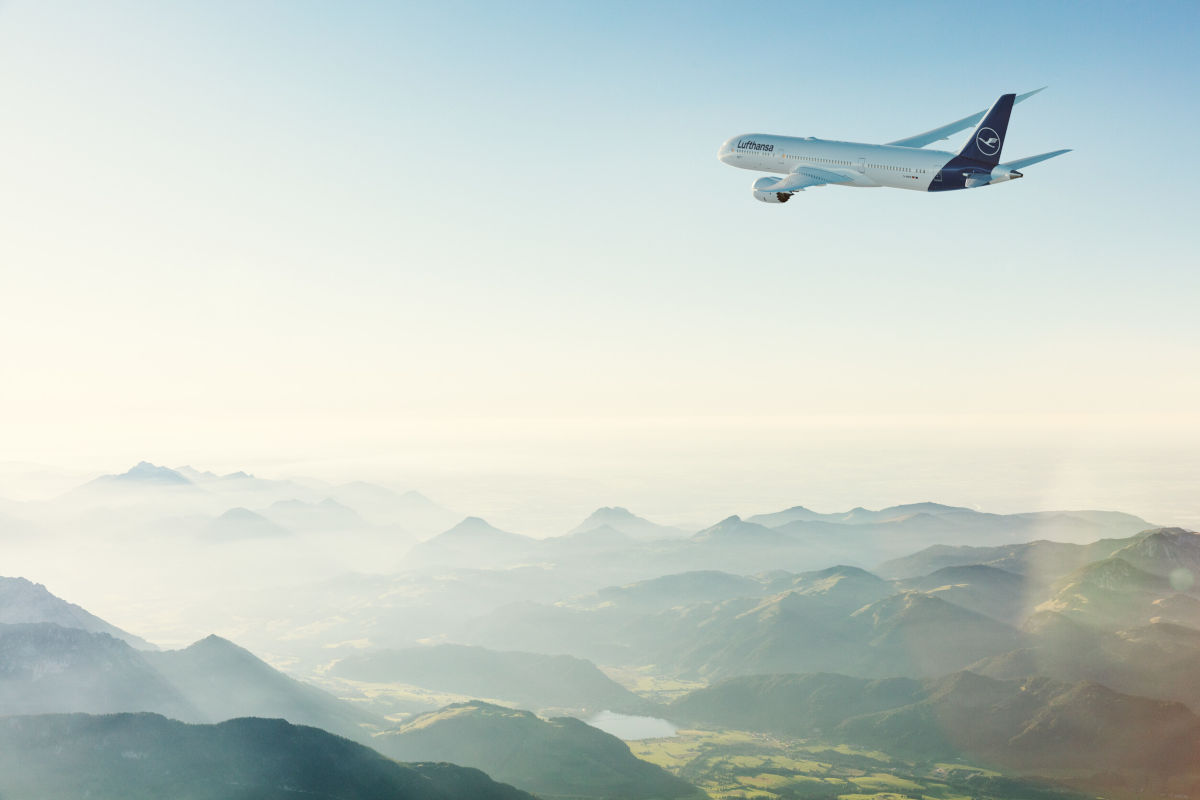 Ein Lufthansa-Flugzeug fliegt in großer Höhe über eine weite, grüne Berglandschaft unter klarem Himmel. Das Panorama zeigt beeindruckende Alpen mit sanften Tälern und einen malerischen See. Die Aufnahme vermittelt das Thema Luftfahrt, Reisen und Natur aus der Vogelperspektive.