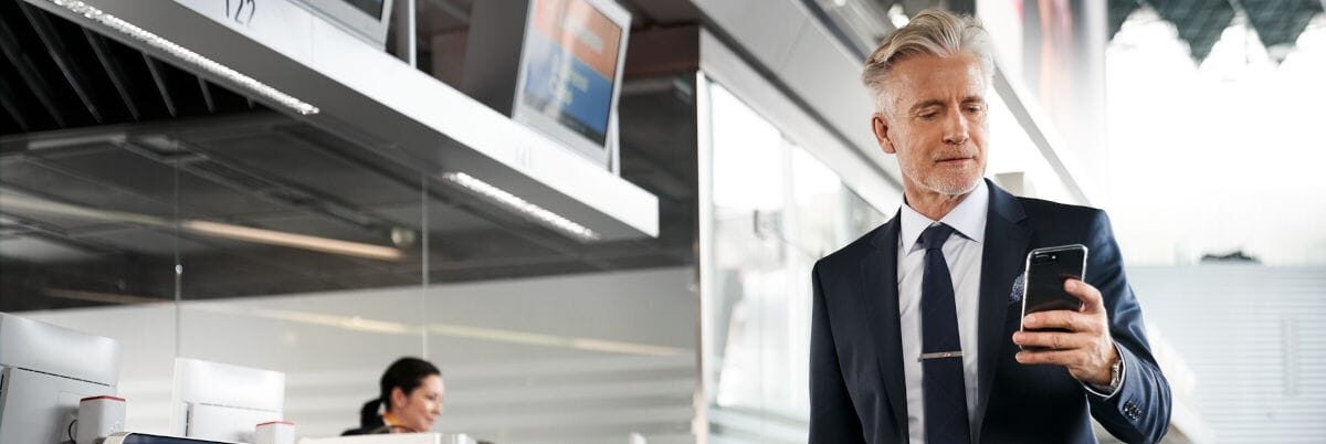Ein Geschäftsmann im Anzug läuft mit seinem Smartphone durch den modernen Lufthansa Check-in Bereich am Flughafen. Im Hintergrund sind weitere Passagiere und Lufthansa-Schalter zu sehen, die das Reisen und Business-Fliegen thematisieren.