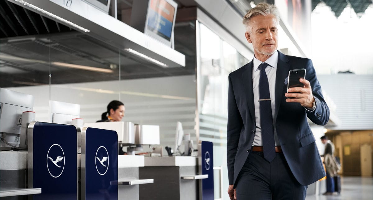 Ein Geschäftsmann im Anzug läuft mit seinem Smartphone durch den modernen Lufthansa Check-in Bereich am Flughafen. Im Hintergrund sind weitere Passagiere und Lufthansa-Schalter zu sehen, die das Reisen und Business-Fliegen thematisieren.