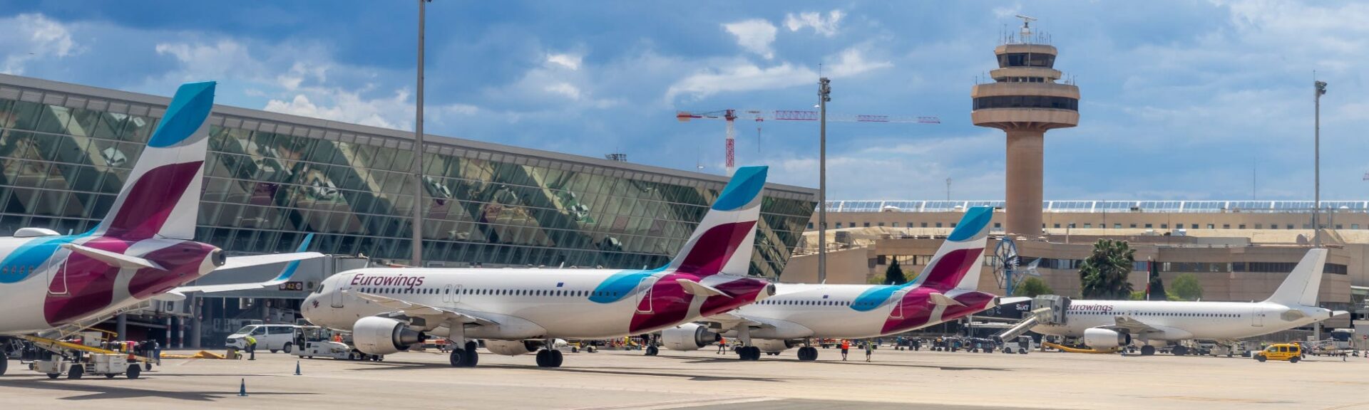 Am Flughafen Palma de Mallorca stehen mehrere Eurowings-Flugzeuge nebeneinander auf dem Rollfeld, im Hintergrund ist das moderne Terminalgebäude und der markante Flughafentower zu sehen. Die Szenerie spielt sich unter einem leicht bewölkten Himmel ab.