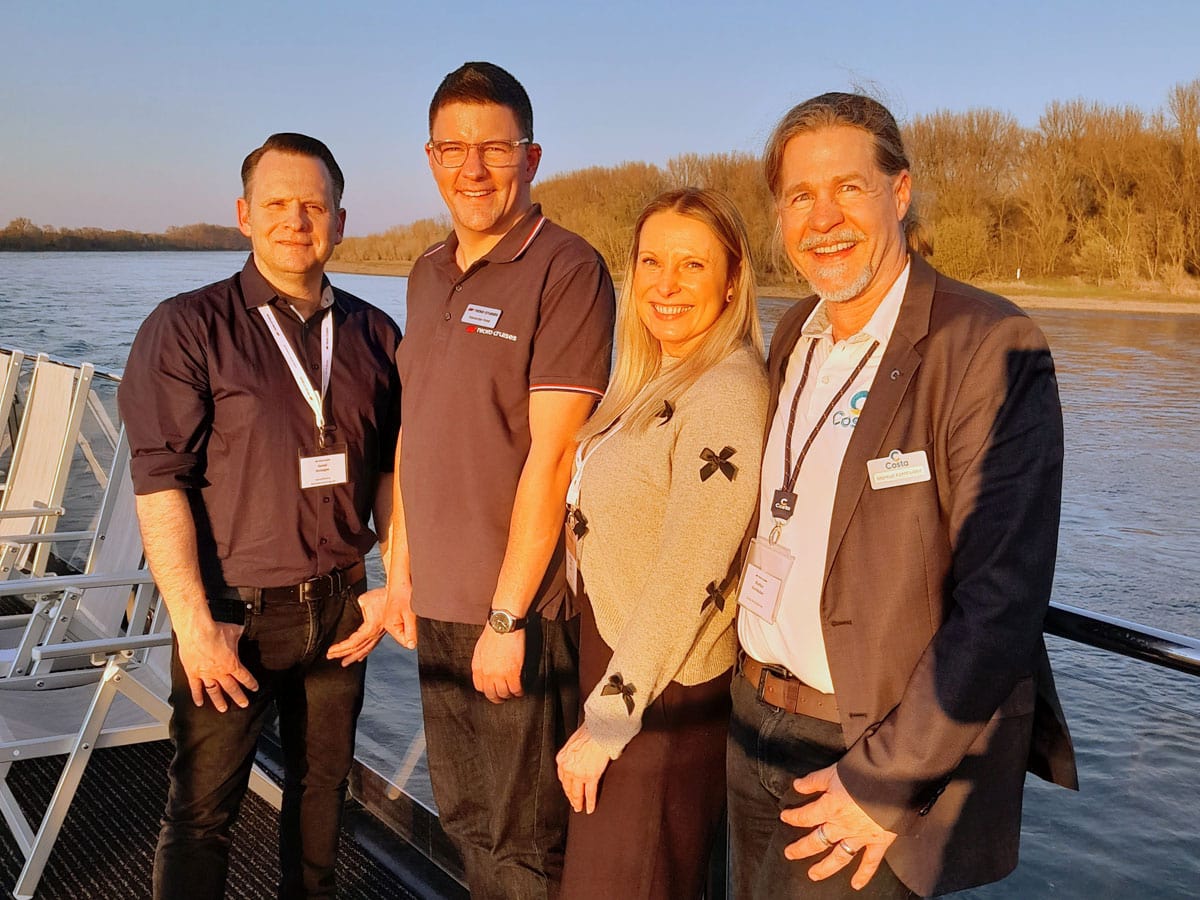 Drei Männer und eine Frau stehen als kleine Gruppe auf dem Sonnendeck eines Flusskreuzfahrtschiffes und blicken lächelnd in die Kamera. De Sonne scheint.