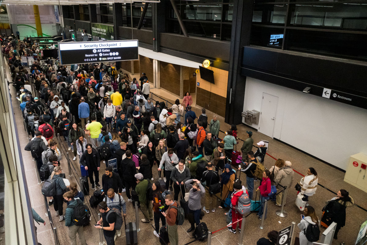 In diesem Foto ist eine lange Warteschlange von Reisenden am Sicherheits-Checkpoint eines Flughafens in den USA zu sehen. Zahlreiche Menschen stehen dicht an dicht mit Gepäck und warten auf die Sicherheitskontrolle. Das Bild zeigt die typische Situation an Flughäfen in den USA mit langen Wartezeiten, vielen Passagieren und geschäftigem Treiben.