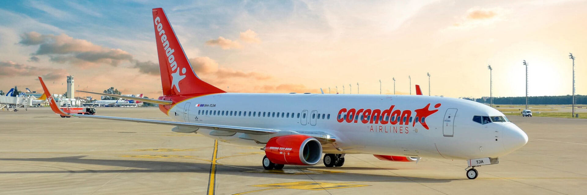 Auf dem Bild ist ein Flugzeug von Corendon Airlines zu sehen, das auf dem Vorfeld eines Flughafens parkt, umgeben von weiteren Flugzeugen und einem Kontrollturm im Hintergrund. Die Maschine vom Typ Boeing 737-800 steht im Mittelpunkt vor einem farbenfrohen Sonnenuntergang.