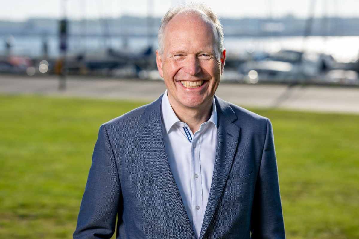Ein freundlich lächelnder, älterer Mann mit kurzem, hellem Haar steht im Freien auf einer grünen Wiese. Er trägt ein hellblaues Hemd und ein dunkelblaues Sakko. Im Hintergrund sind unscharf ein Gewässer und mehrere Boote zu erkennen, was auf einen Hafen oder eine Uferpromenade hindeutet.