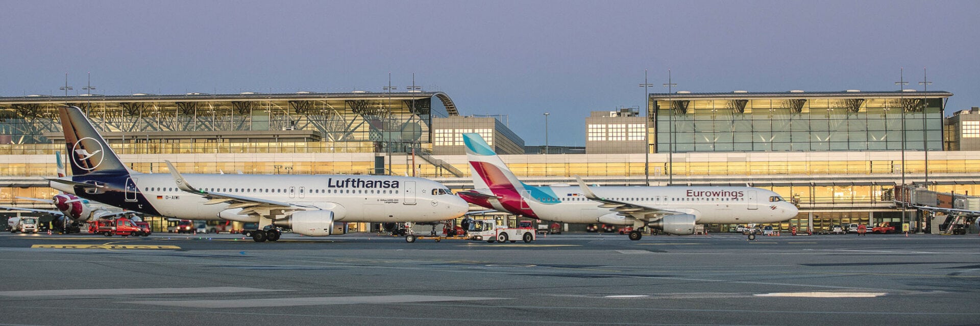 Zwei Flugzeuge der Airlines Lufthansa und Eurowings stehen nebeneinander auf dem Rollfeld eines modernen Flughafens, im Hintergrund sieht man ein gläsernes Terminalgebäude. Die Maschinen sind bereit für den Abflug, während Fahrzeuge und Personal auf dem Vorfeld arbeiten.