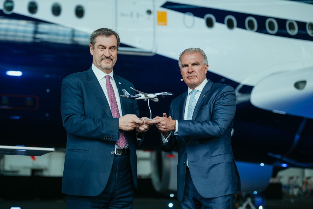 Zwei Männer in Anzügen stehen vor einem Lufthansa-Flugzeug und halten gemeinsam ein Modellflugzeug in den Händen. Die Szene spielt in einem Hangar am Flughafen München und symbolisiert eine feierliche Übergabe oder Präsentation.