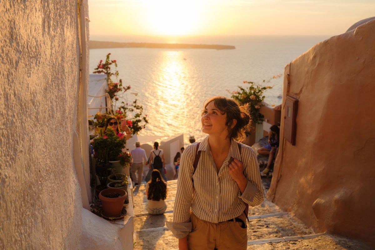 Eine junge Frau mit Rucksack geht eine schmale Treppe zwischen weiß getünchten Häusern hinauf und blickt lächelnd nach oben. Im Hintergrund glitzert das Meer im warmen Licht eines Sonnenuntergangs, während mehrere Menschen die Stufen hinuntergehen oder sitzen; Blumen in Töpfen säumen den Weg.