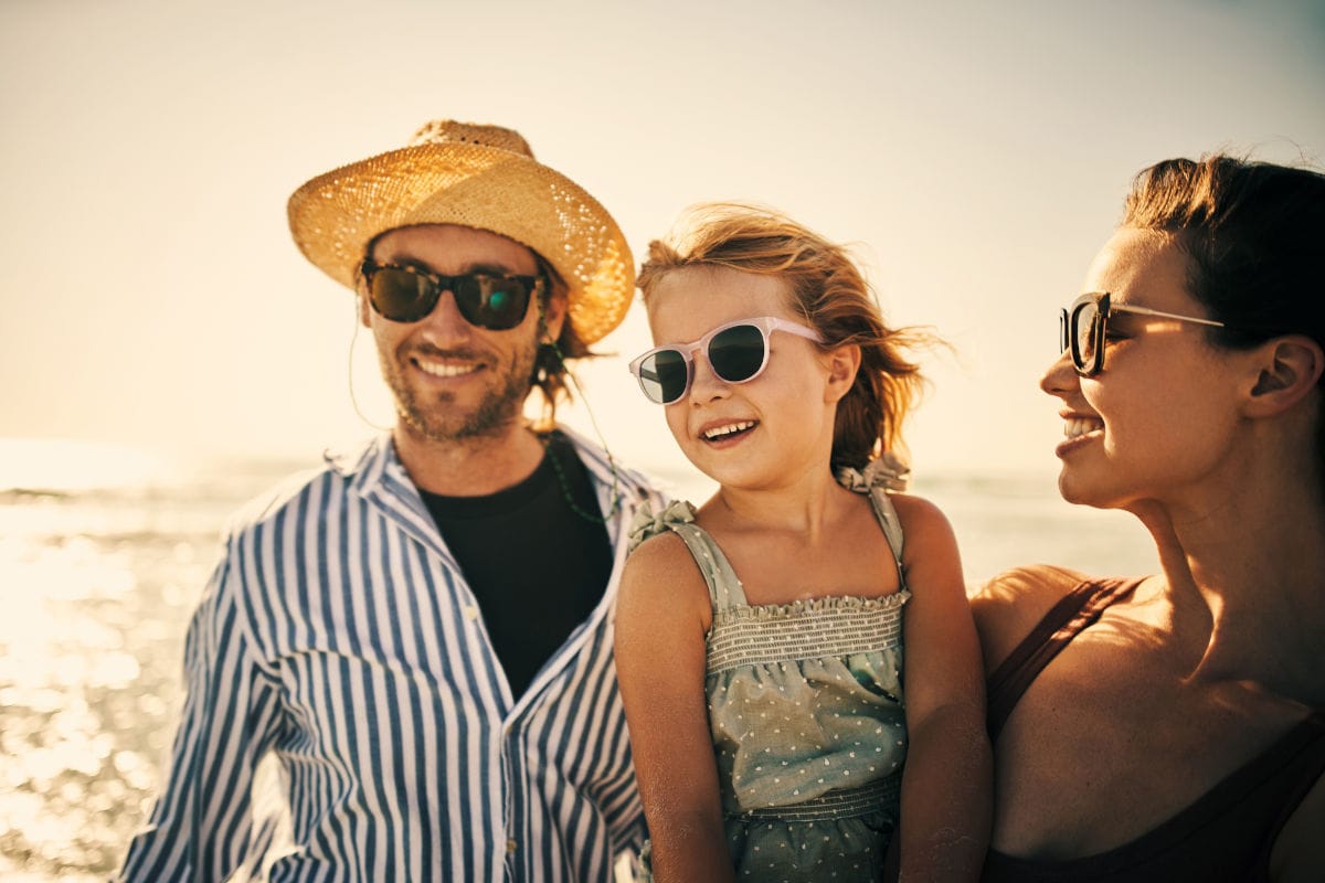 Eine Familie am Strand im warmen Abendlicht: Ein Mann mit Strohhut und Sonnenbrille steht neben einer Frau, die eine lächelnde kleine Tochter auf dem Arm hält; alle tragen Sommerkleidung und schauen entspannt in die Ferne, während im Hintergrund das Meer glitzert.