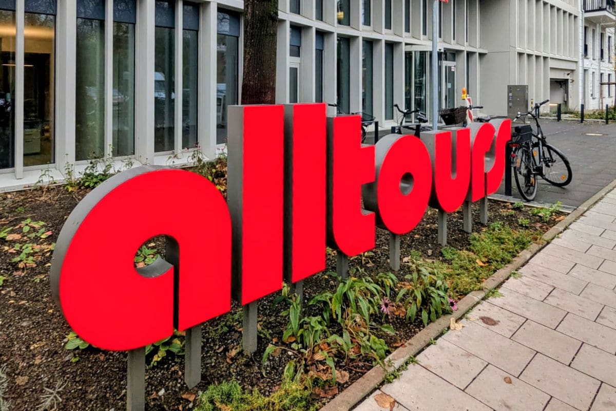 Rotes „alltours“-Logo als große freistehende Buchstaben vor einem modernen Bürogebäude. Daneben stehen Fahrräder, und im Vordergrund befinden sich Pflanzen entlang eines Gehwegs.