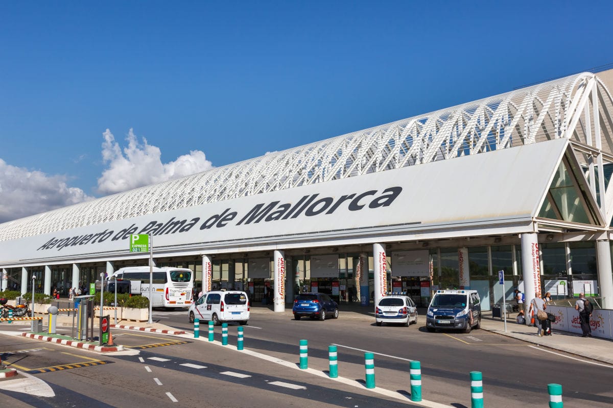 Das Bild zeigt den Eingangsbereich des Flughafens Palma de Mallorca mit dem modernen Terminalgebäude, mehreren parkenden Taxis und Bussen sowie Passagieren vor dem Eingang. Die Fassade trägt deutlich sichtbar den Schriftzug „Aeropuerto de Palma de Mallorca“.